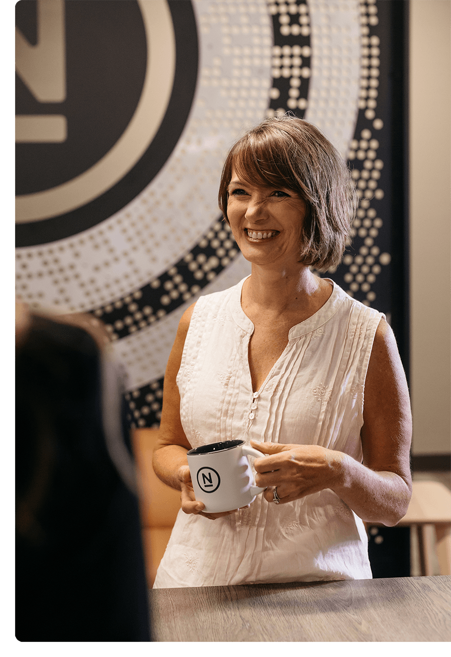 Woman with short brown hair smiling, holding a coffee cup, talking to coworkers