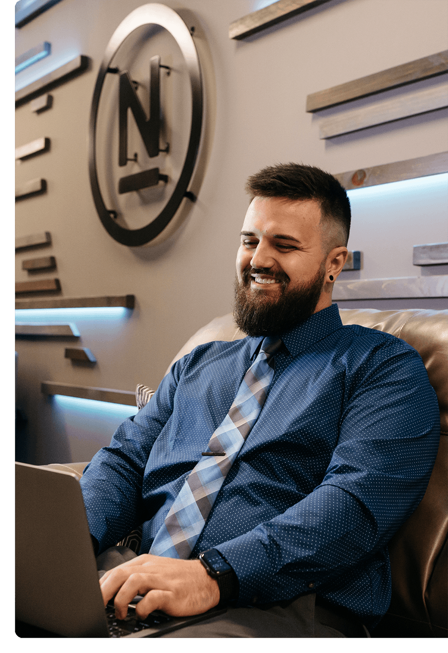 Man with a beard in a dress shirt and tie smiling while working on a laptop.