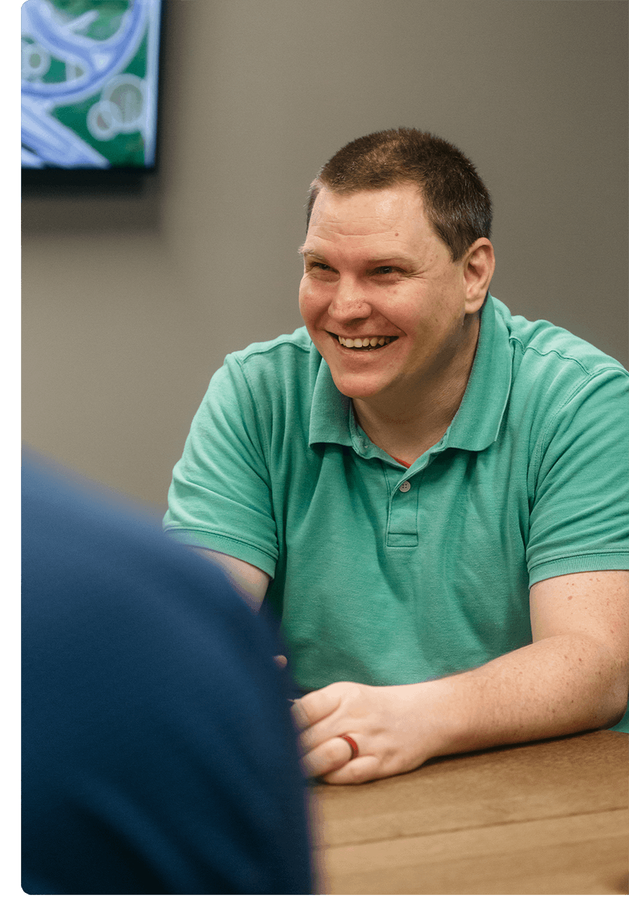 Man in teal shirt smiles and laughs while sitting at a lunch table with coworkers