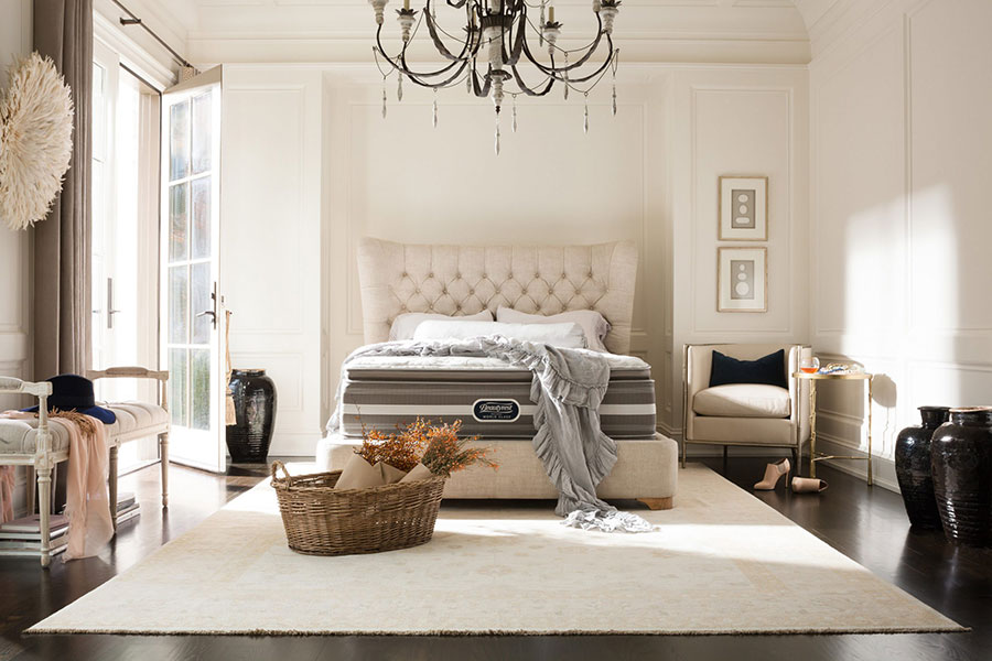 A large bedroom with beige walls, a beige carpet, and beige bed. The bed has a Beautyrest mattress on it.