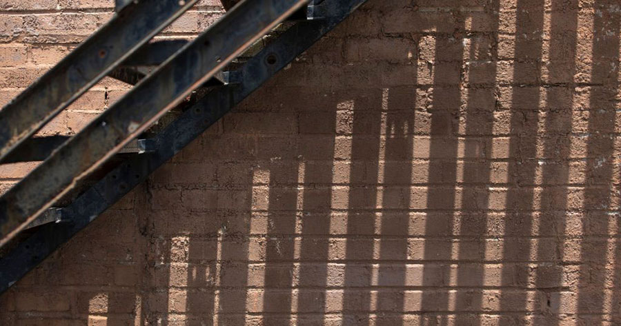 An abstract image of a brown brick wall. A metal staircase connected to the wall casts shadow lines down the wall.