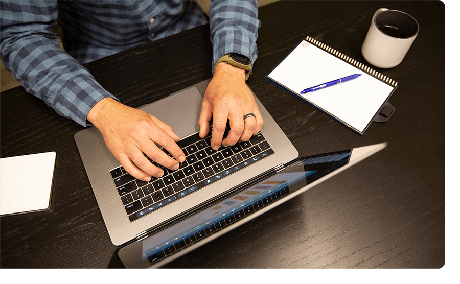 Man working on a laptop with a notepad and coffee cup beside him.