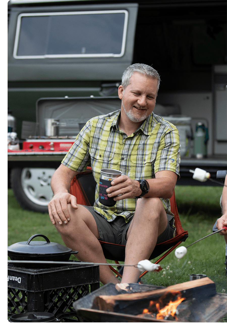 Man with gray hair and goatee sits at a campsite holding his coffee.