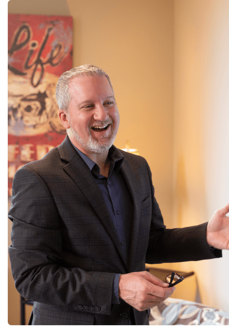 Man with white hair and goatee talks and laughs. He is wearing a black blazer.