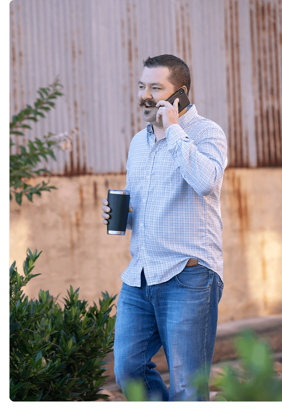 Man with large mustache in blue long-sleeve shirt, holding coffee, stands and talks on his cell phone.