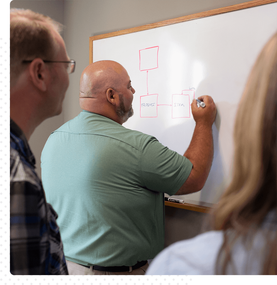 A man in a teal shirt writes on a whiteboard as two coworkers look on.