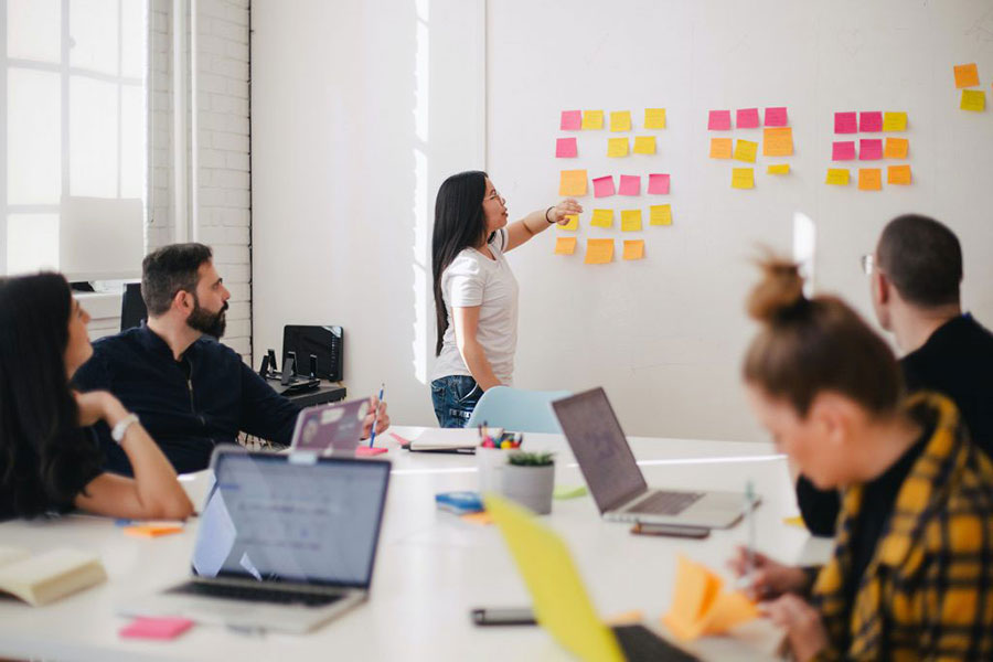 A woman stands in front of a meeting presenting a project plan on a whiteboard.
