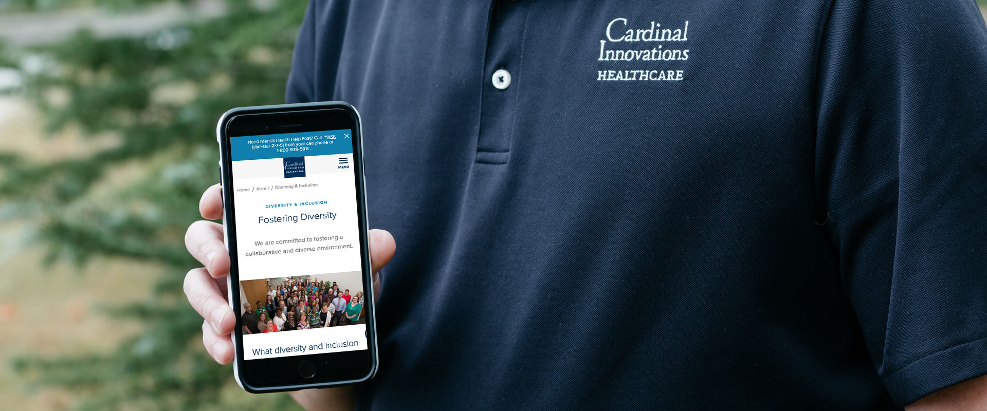 A person is holding a phone showing the "Fostering Diversity" page of the Cardinal Innovations Healthcare website, while wearing a branded polo shirt.