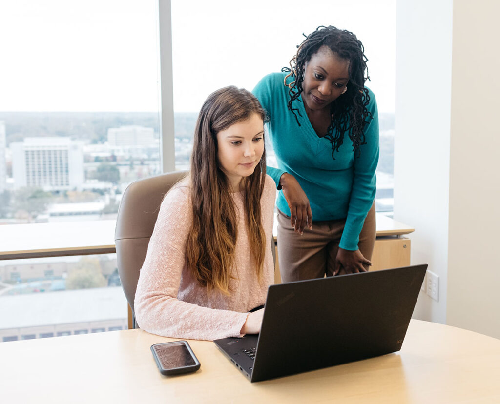 A photo of two women in an office, one standing behind the other, both looking at a laptop screen.