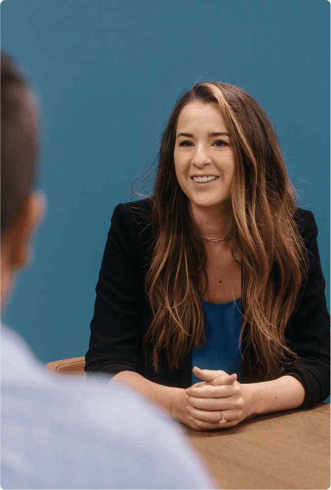 linkedin-sales-navigator-hrhjn6ZTgrM-unsplash 1(3) Woman sitting at a table smiling. In the foreground, an out-of-focus man, who she appears to be talking to.