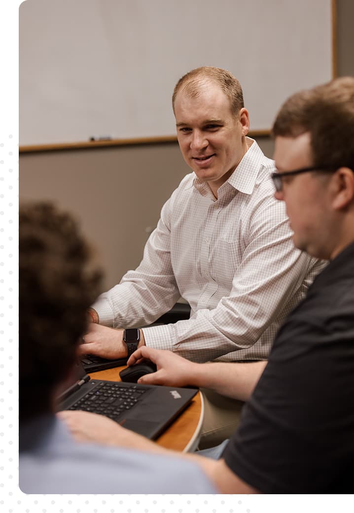 Two men sitting at a table with computers. One is working and one is smiling, looking off in the distance.