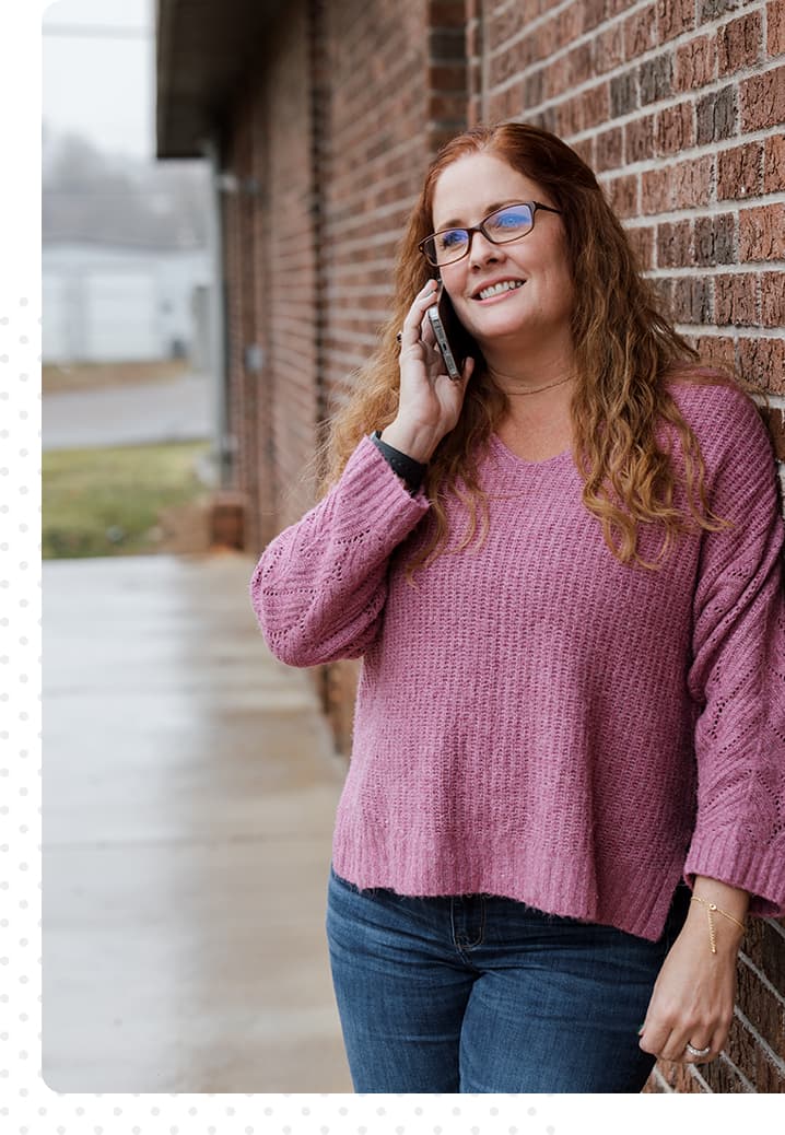 Image of a red haired woman with a pink sweater and jeans talking on the phone.