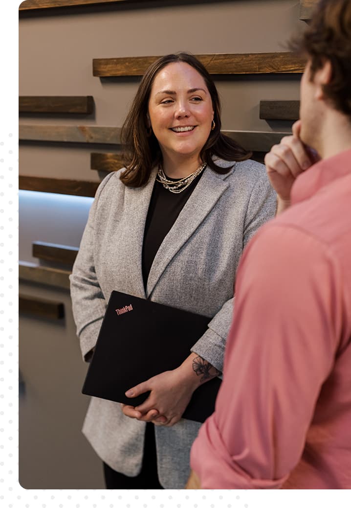 A man and woman talking. She is smiling, holding a computer, and he has his hand on his chin.