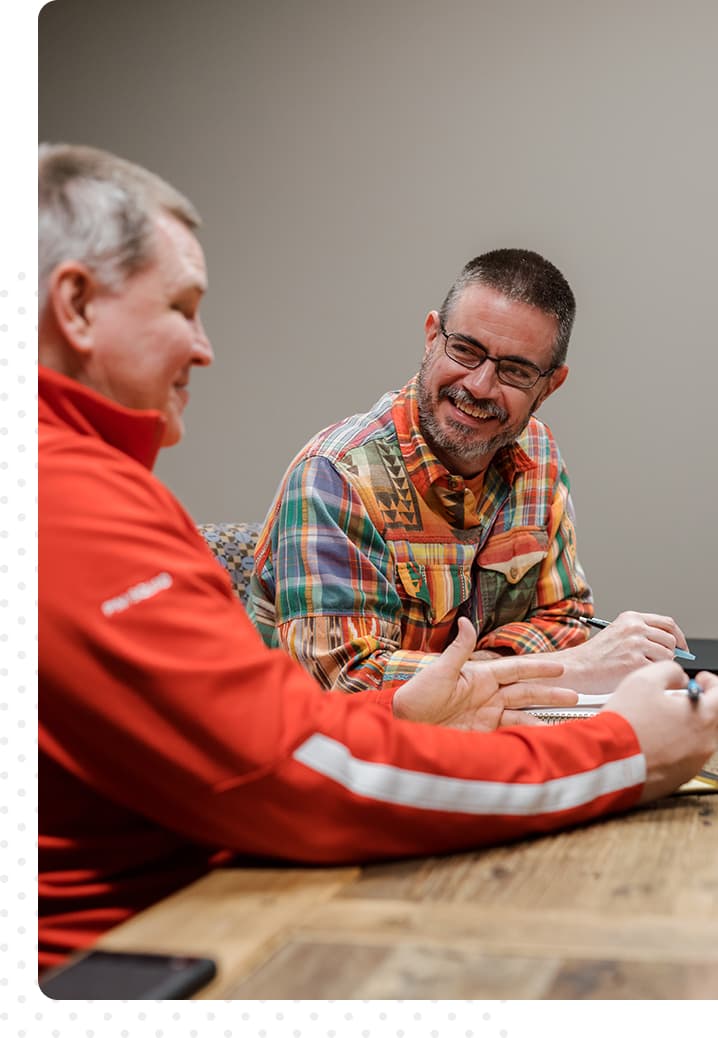 Two men sitting at a table. On the left, a man in red smiles and looks at his notebook. On the right, a man in colorful plaid smiles at the man on the left.