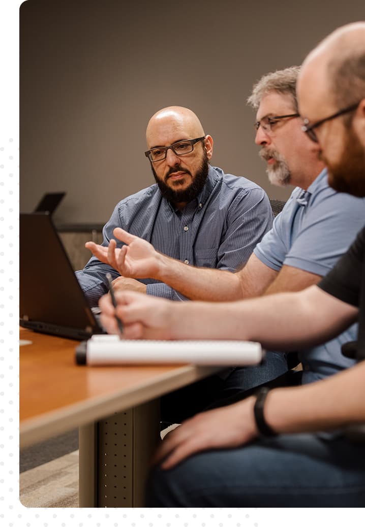 Three men sitting at a table, looking at the same laptop, having a conversation