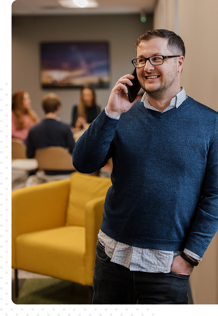 Image of a man in a blue shirt and glasses talking on the phone, smiling, with other people sitting in the background.