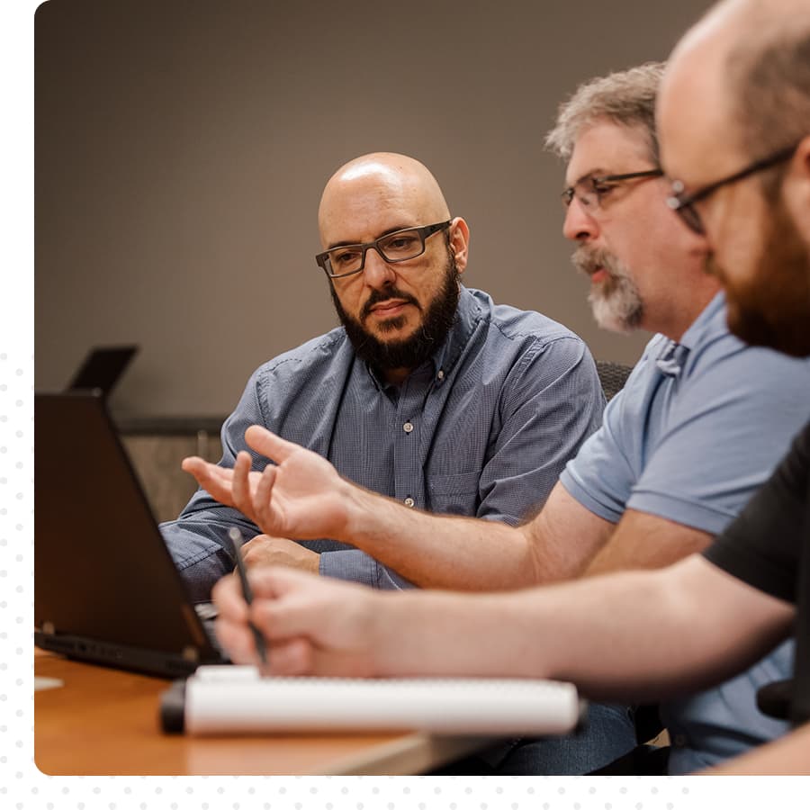 Three men sit at a table, looking at a computer and talking. 