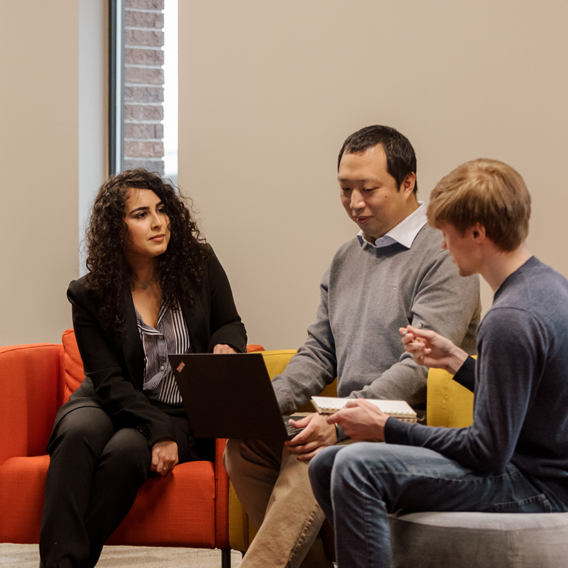 A woman in a black suit and two men sit on colorful chairs in a casual meeting. One man is holding a laptop, while the other is writing in a notebook.