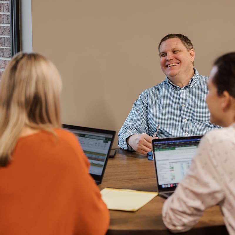 A man smiles while sitting at a table with two women. Laptops and documents are placed on the table.
