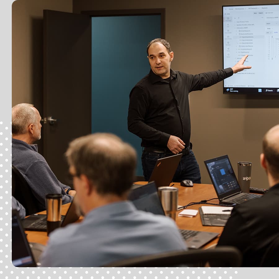 Man leading a group discussion. He is dressed in black, gesturing toward a screen at the front of the room, talking to three other men sitting at a table with their computers and coffee mugs.