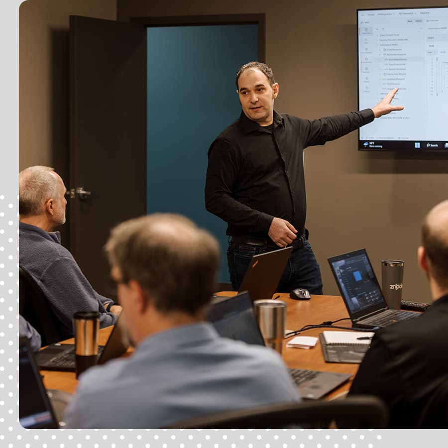 Aaron Jestrab presentation Man leading a group discussion. He is dressed in black, gesturing toward a screen at the front of the room, talking to three other men sitting at a table with their computers and coffee mugs.