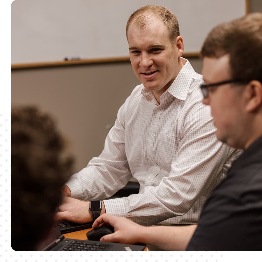 Three men sitting at a table having a conversation. One is smiling. Another is looking at a computer. The third is not fully in frame.