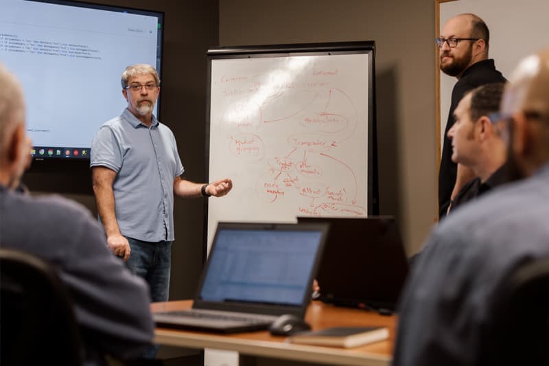 Man at whiteboard, sketching out a PIM flow, with other men surrounding him in discussion.