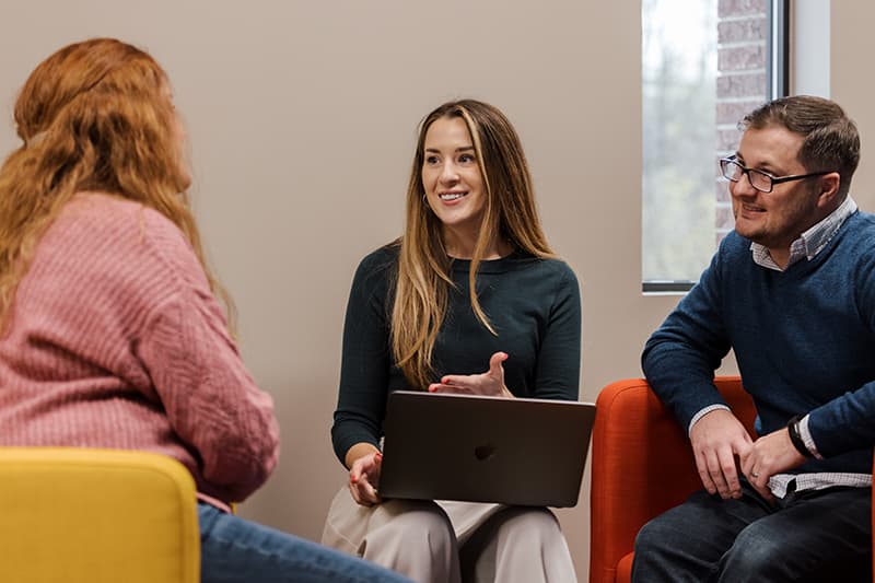 Image of two women and one man seated. One woman is holding a computer and smiling.