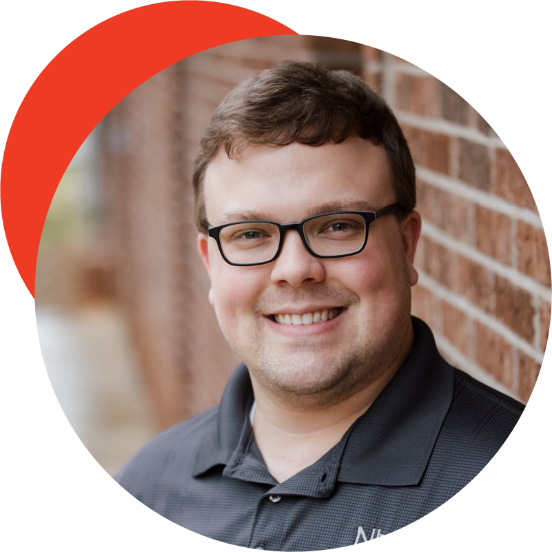 Headshot of Zachary Helbert. Man with brown hair, glasses, and gray polo with brick in the background.