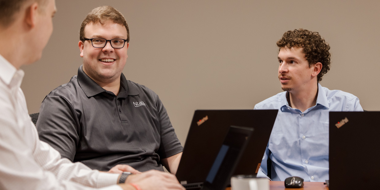 Image of three men. On the left, a man in a white button-down top. He is looking at the second man, so you can only see the right side of his face. In the center, a man with a gray shirt and glasses sits in front of his laptop. On the right, another man with curly hair and a blue button down looks at the man in the center.