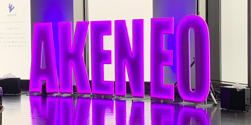 Large purple letters lit internally with lights spell out Akeneo. Lights are in front of tall windows. 