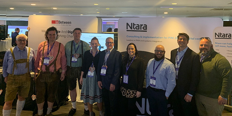 Nine people stand smiling, side-by-side. On the left, three men and one woman represent the InBetween publishing software team. They are dressed in traditional German clothing. On the right, four men and one woman represent the Ntara systems integrator team. Behind each team is a large sign that has its company logo.  