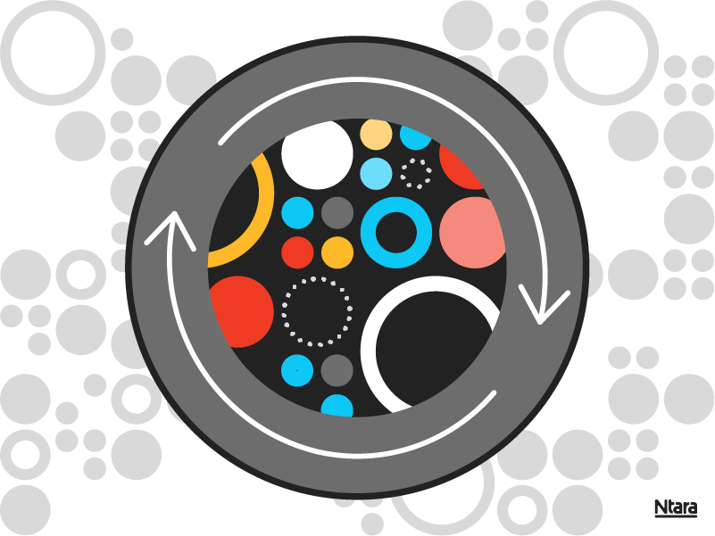 Grey circle with a black outline. At the center, a black circle filled with other circles in various colors, shapes, and sizes. On the grey of the outer circle are two white arrows, indicating motion.